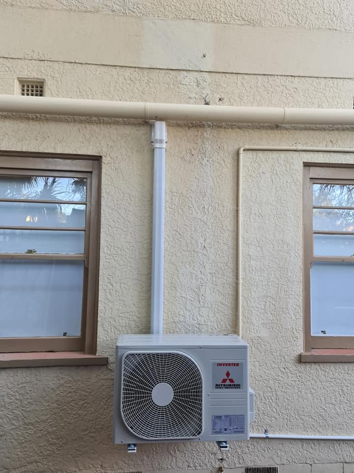 A Mitsubishi outdoor unit mounted on a rendered wall, with a white pipe cover running up the wall.
